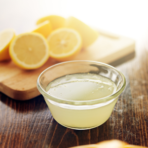 A small bowl of lemon juice sits on a wooden table in front of some sliced lemons. 