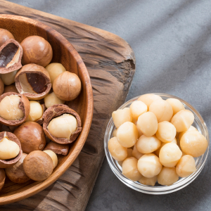 A glass bowl filled with unshelled macadamia nuts next to a wooden bowl filled with macadamia nuts with shells on a grey background