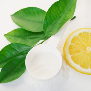 A white plastic scoop filled with ascorbic acid powder next to a lemon slice and sprig of leaves