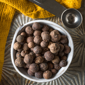 White bowl filled with whole allspice next to a metal measuring spoon and a yellow towel