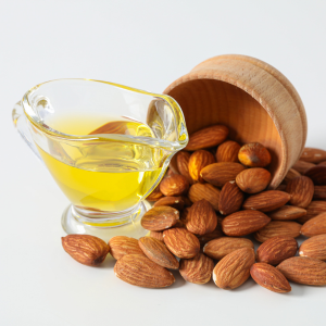 Sweet almond oil in a glass container next to almonds and a wooden bowl with a white background