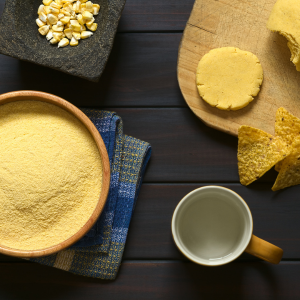 A wooden bowl of cornmeal next to a mug, tortilla chips, cornbread dough, and a bowl of kernels on a dark wooden background