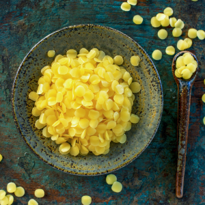 A bowl and spoon filled with unrefined beeswax pellets with a blue textured background