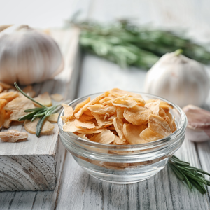 Glass bowl filled with garlic flakes surrounded by a wooden background with rosemary and garlic