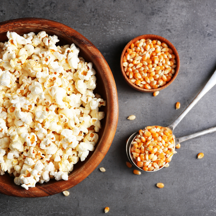 Popped popcorn in a wooden bowl next to popcorn kernels in a metal spoon and wooden bowl on a grey surface