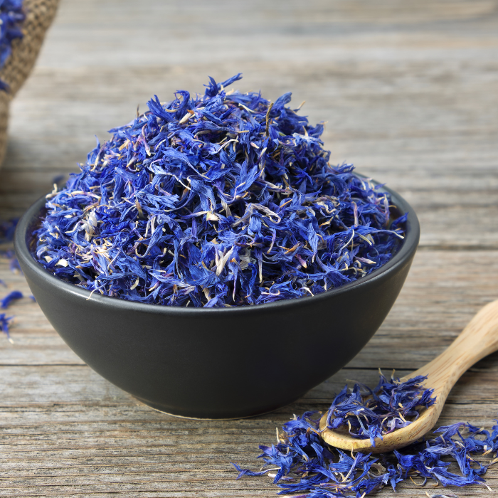 Blue cornflower in a black bowl next to a wooden spoon on a grey wooden background