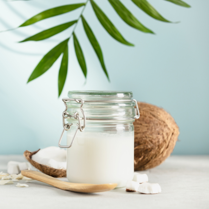 Coconut oil in a glass jar next to coconut pieces and a wooden spoon with a light blue background and a leaf