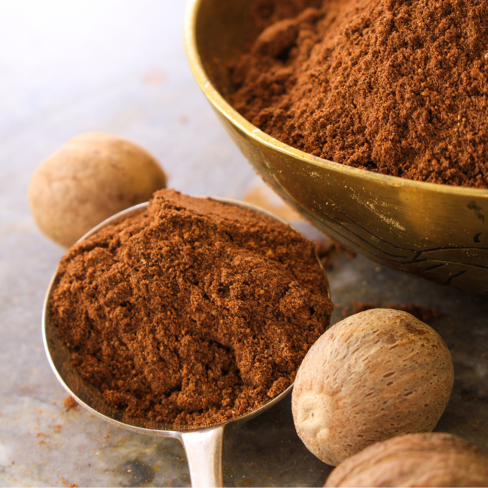 Ground nutmeg in a metal bowl and spoon next to whole nutmeg