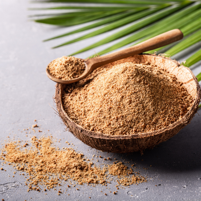 Coconut sugar in a coconut shell next to a palm leaf and wooden spoon on a grey surface