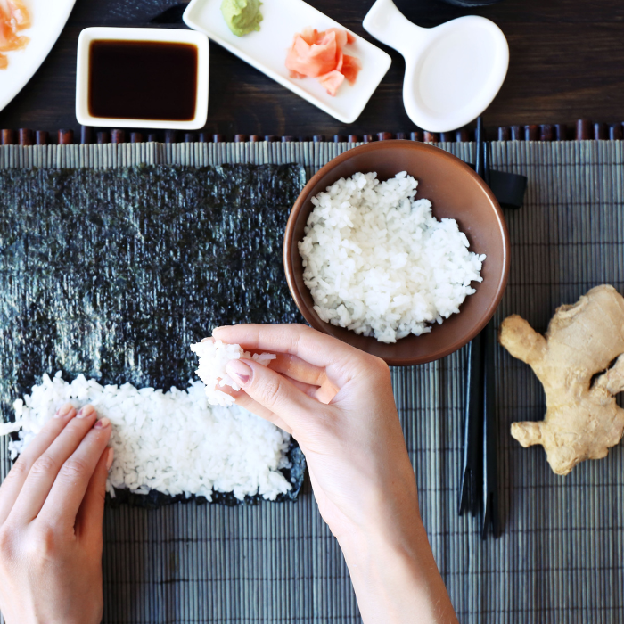 Cooked sushi rice being layed out on seaweed with ginger, wasabi, and soy sauce on a sushi mat