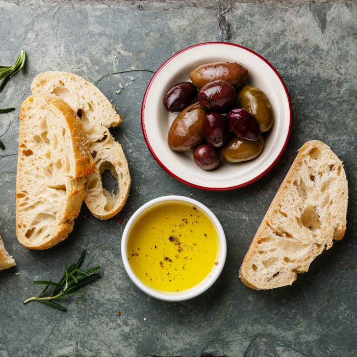 Olive oil in a white bowl next to a bowl of olives, slices of bread, and rosemary sprigs on a stone surface