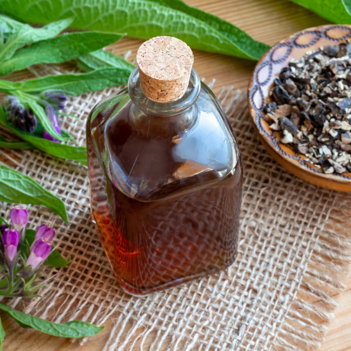 A glass bottle filled with comfrey root infused oil next to a wooden bowl of comfrey rood on a wooden background