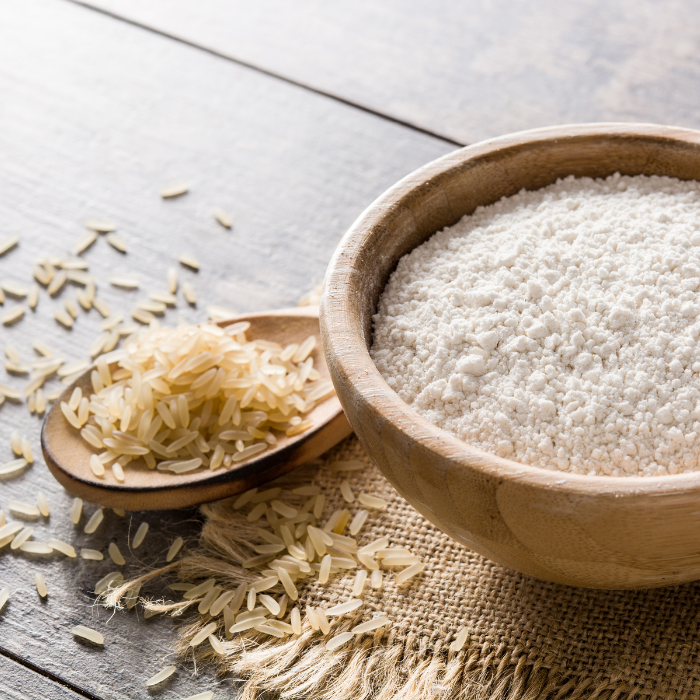 Brown rice flour in a white bowl next t o a wooden spoon of brown rice on burlap fabric