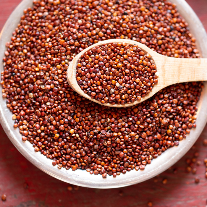 A wooden bowl filled with red quinoa with a wooden spoon and a red wooden background