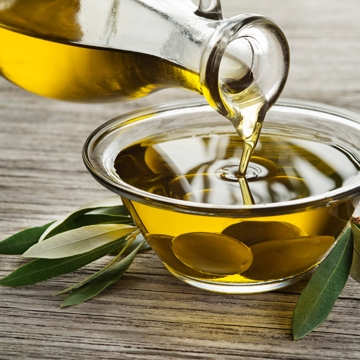 Olive oil being poured into a glass bowl with olives and olive leaves on a wooden surface