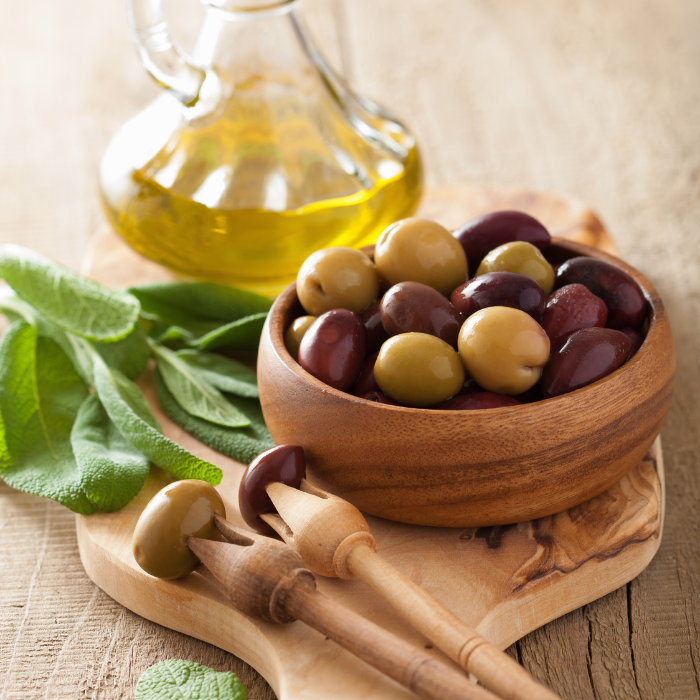 A wooden bowl filled with olives next to sage leaves and a bottle of olive oil on a wooden surface