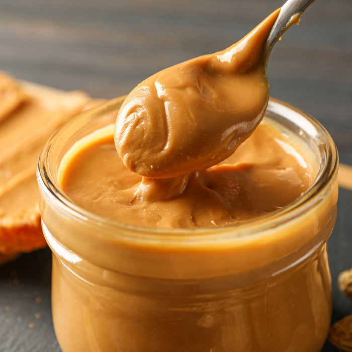 Smooth peanut butter in a glass jar with a metal spoon on a grey surface