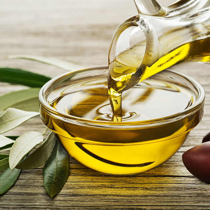 Olive oil being poured into a small glass bowl next to kalamata olives and leaves on a wooden surface