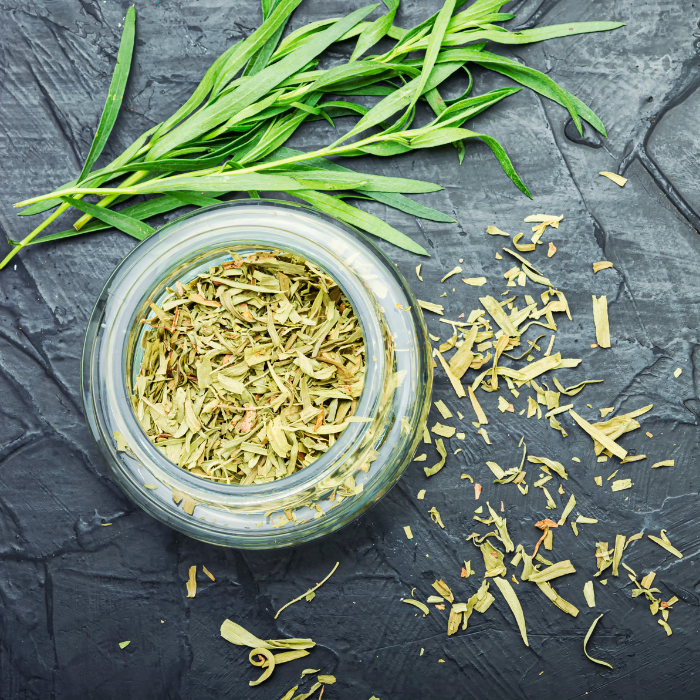 Dried tarragon in a clear glass jar next to fresh tarragon on a black textured background