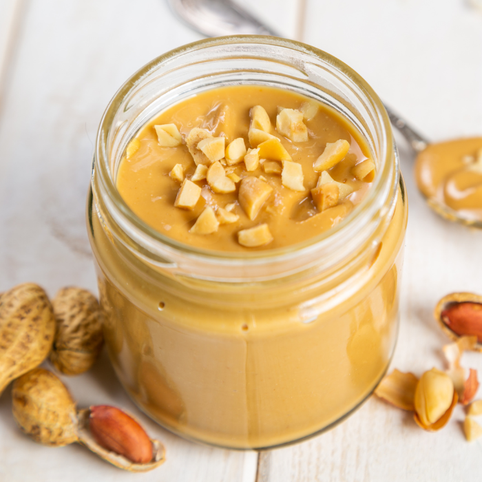 Crunchy peanut butter in a glass jar next to peanuts on a white wooden surface