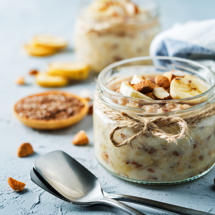 Oatmeal in a clear glass jar with a twine bow, banana, and almond pieces on a blue painted surface with metal spoons