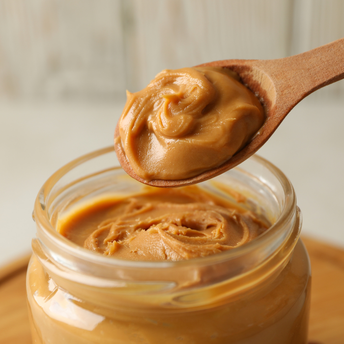 Smooth peanut butter in a clear glass jar with a wooden spoon on a wooden dish