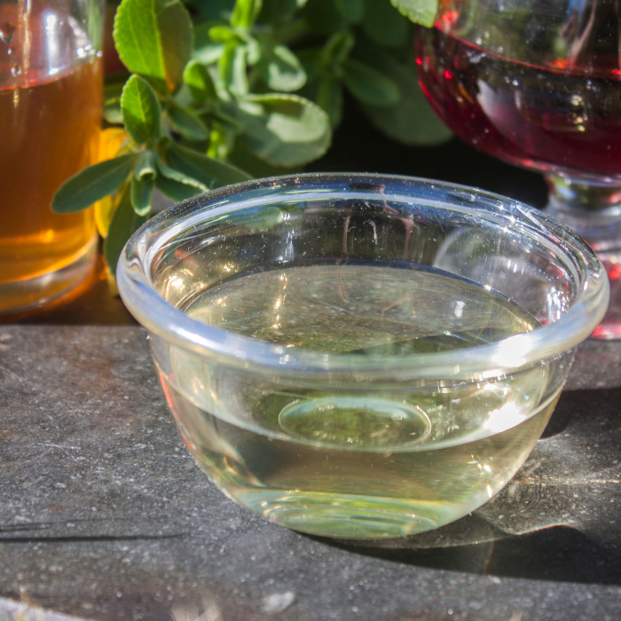 White vinegar in a glass bowl next to herbs and glasses of other types of vinegar on a stone surface