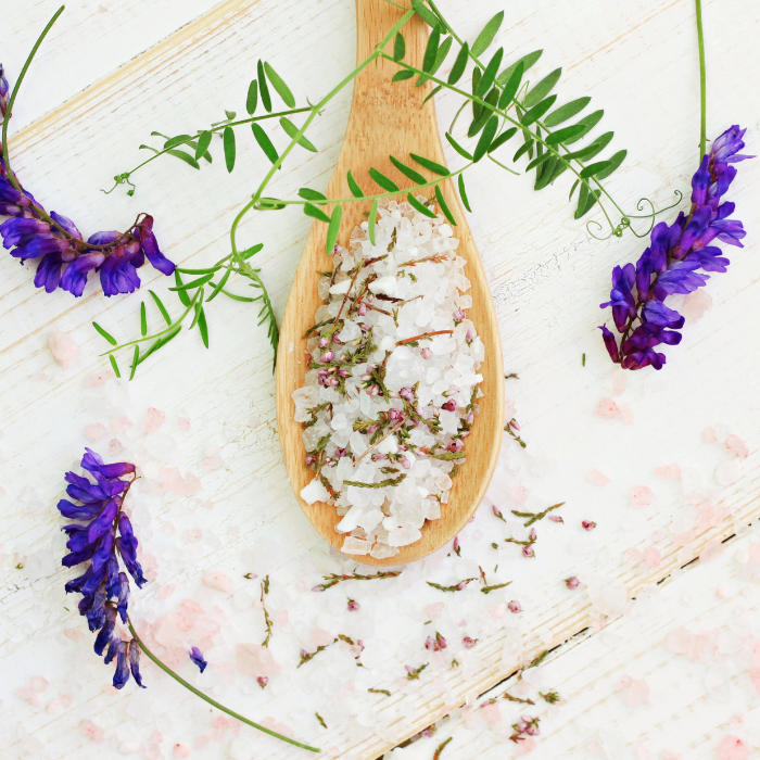 Epsom salt mixed with bath herbs in a wooden spoon next to herbs and flowers on a white wooden surface