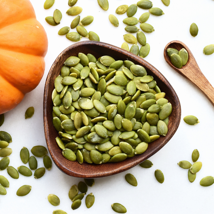 Raw pumpkin seeds in a wooden bowl next to a pumpkin and wooden spoon on a white background