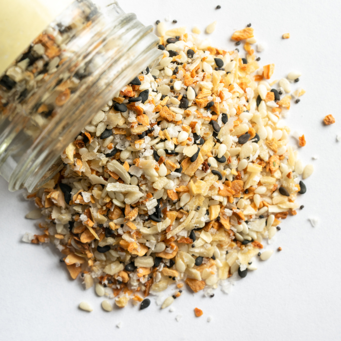 Everything bagel seasoning spilling out of a clear glass jar onto a white background