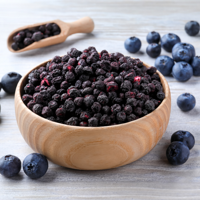 Unsulphured dried blueberries in a wooden bowl next to a wooden spoon and fresh blueberries