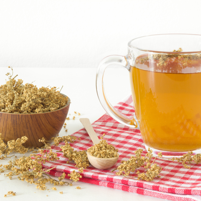 Dried elder flowers being steeped as tea in a clear mug next to a wooden bowl and spoon filled with dried elder flowers