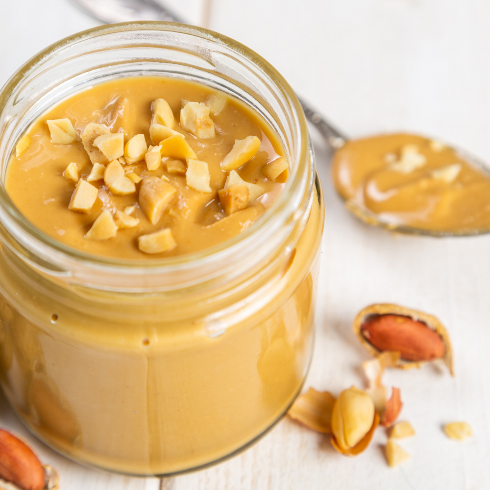 Peanut butter in a glass jar with peanut pieces on a white wooden background