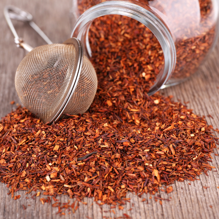 Red rooibos tea coming out of a clear glass jar next to a pair of mesh tea tongs on a wooden background