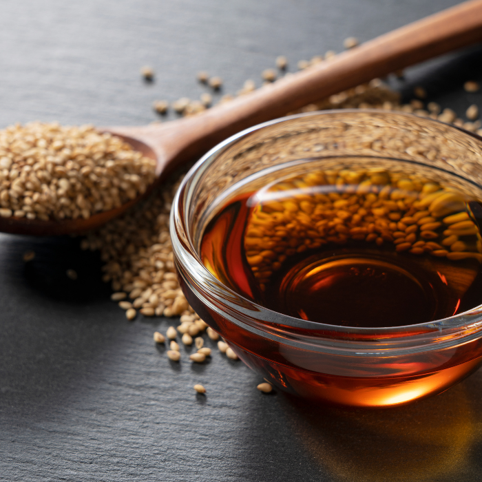 Organic sesame oil in a glass bowl next  to a wooden spoon filled with sesame seeds